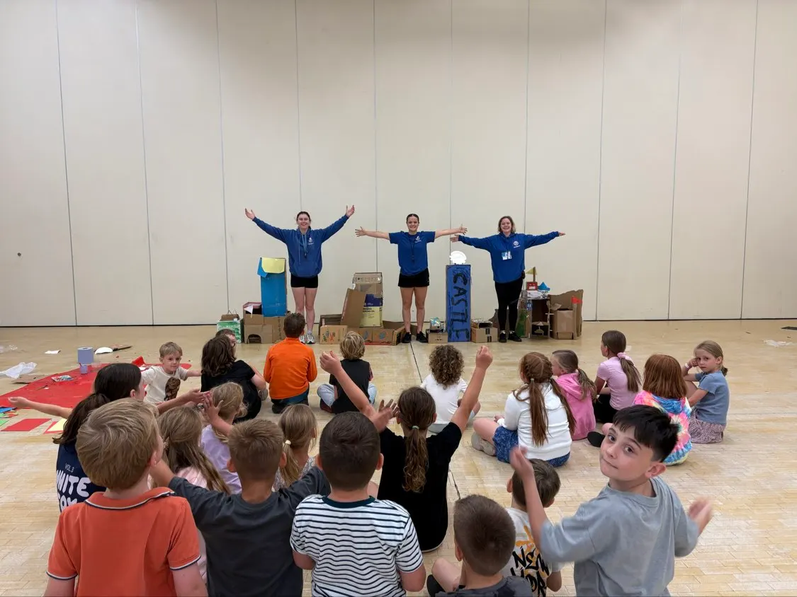 Three instructors in blue uniforms stand on a stage made of cardboard boxes with arms raised, entertaining a group of seated children in a sports hall.