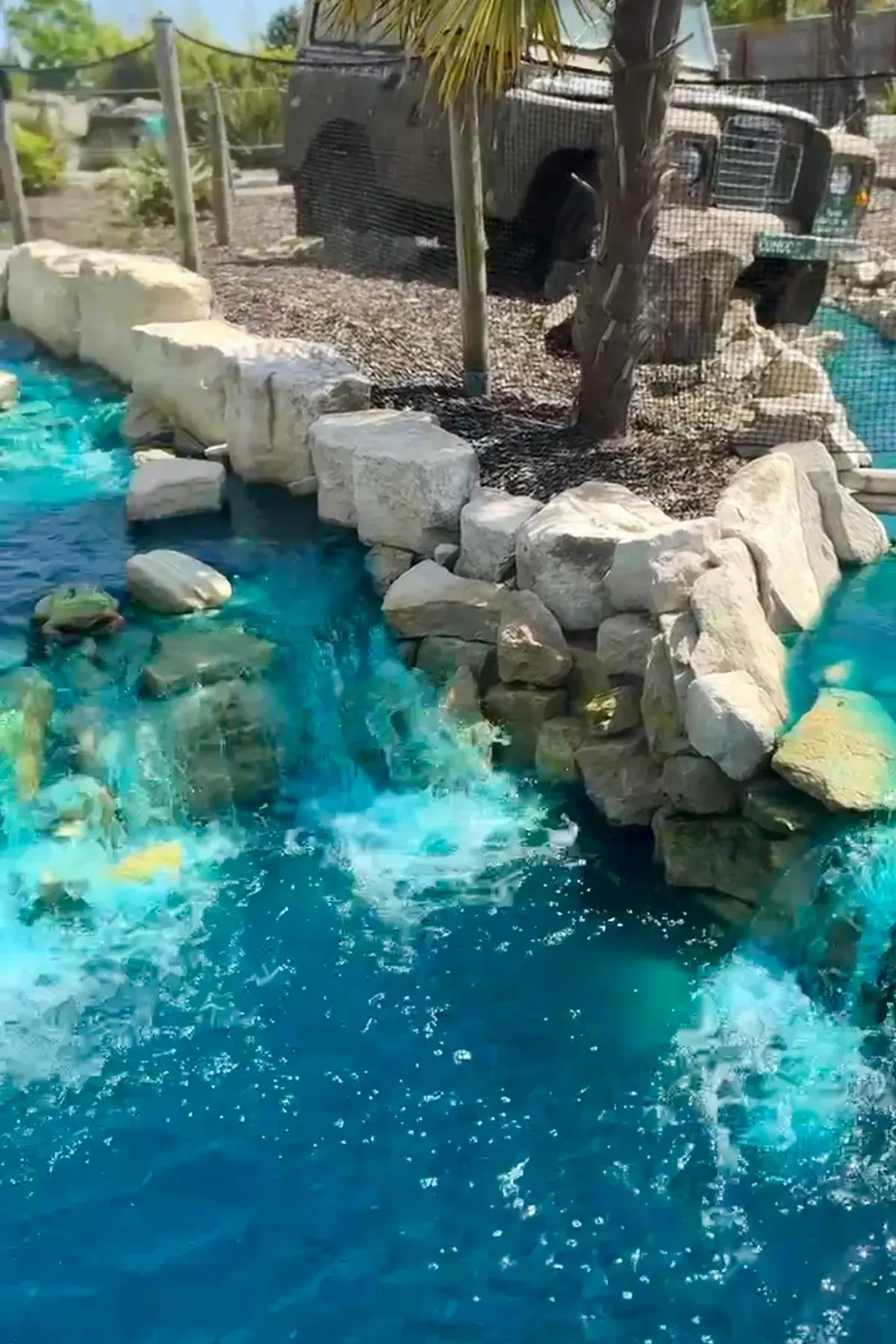 Tropical-themed miniature golf course featuring bright turquoise water, white rocks, and palm trees beside an enclosure.