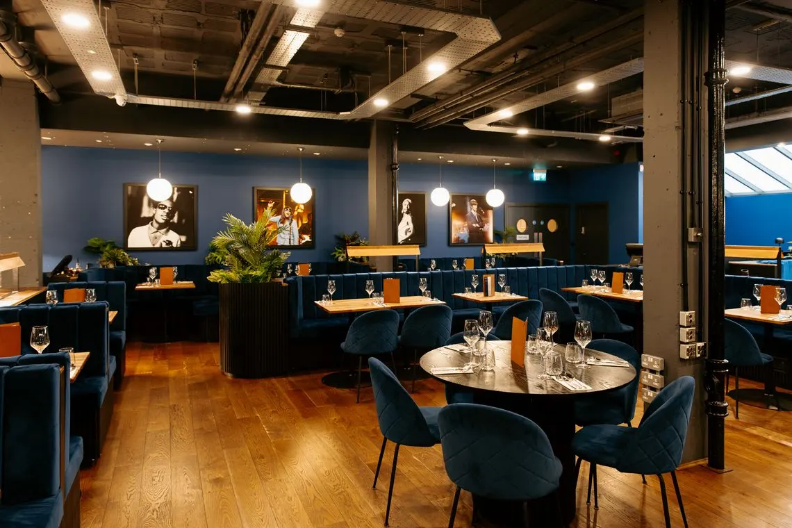 Contemporary restaurant interior with dark blue banquette seating, round tables set for dining, and portrait artwork on walls.