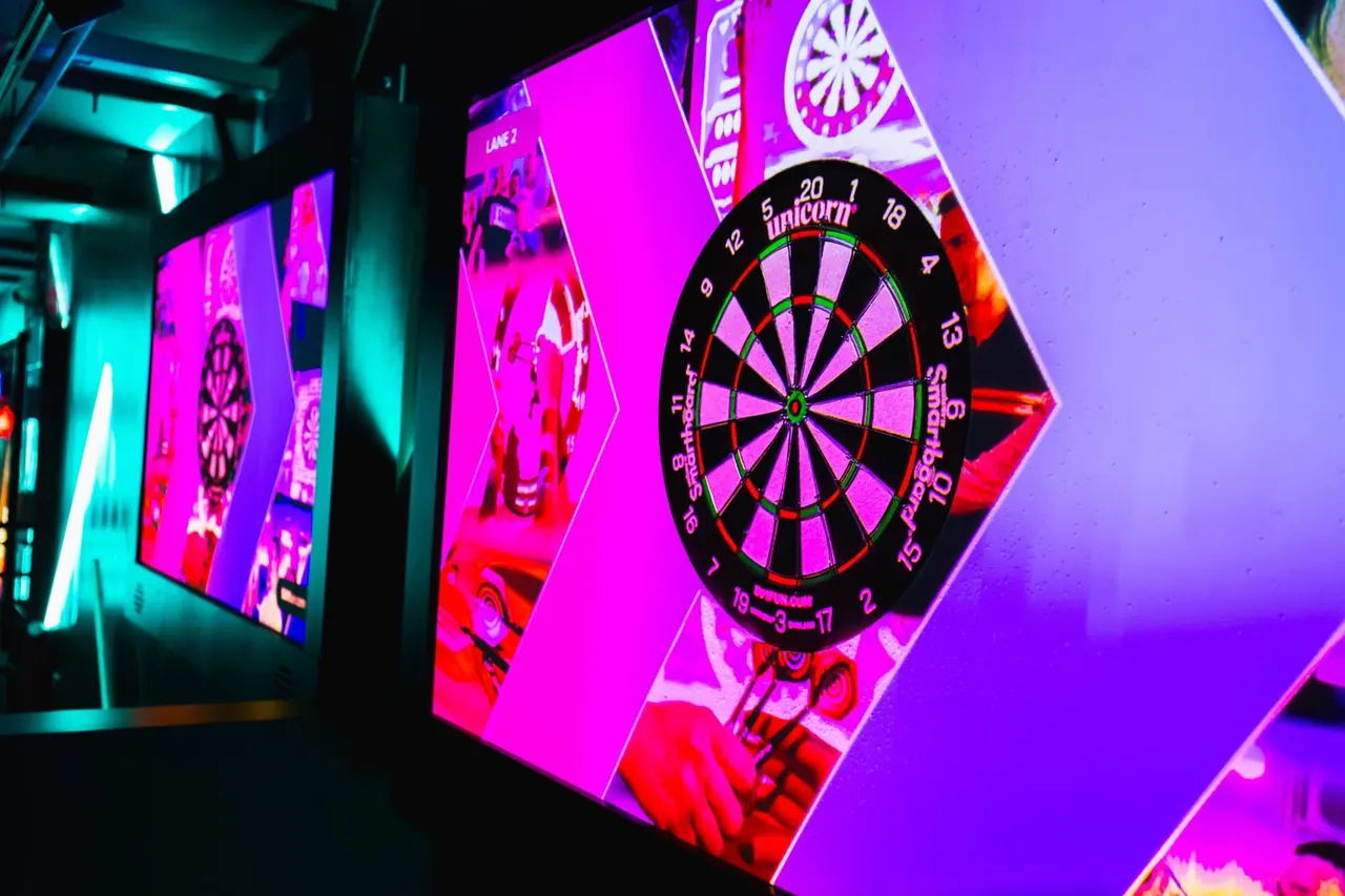 Electronic dartboards illuminated in bright magenta and cyan neon lighting in a modern entertainment venue.