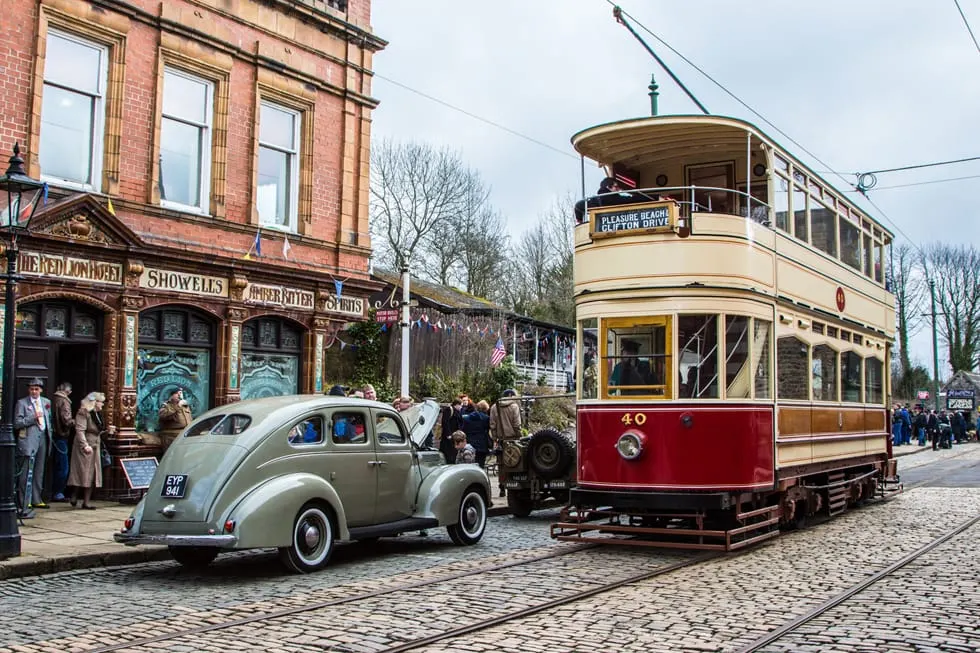 Vintage tram and classic car on cobbled street at heritage transport museum, with period buildings and visitors.