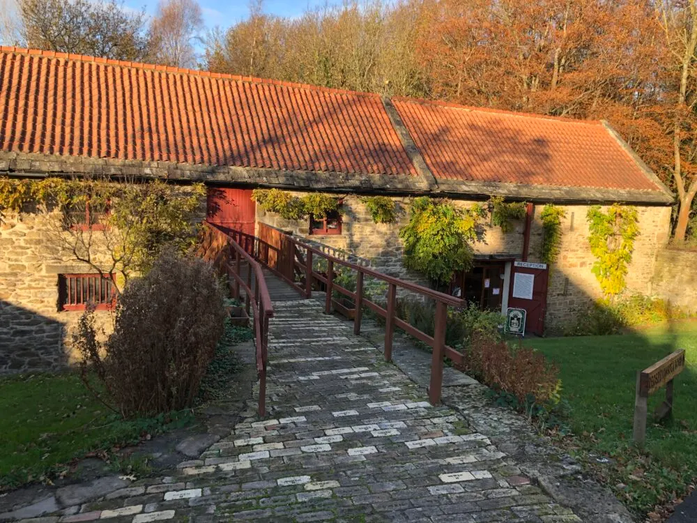 Stone building with terracotta roof and wooden ramp leading to entrance, autumn trees in background.