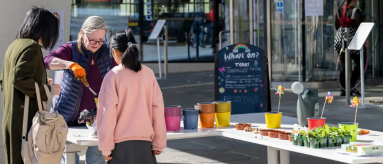 Adults and children at an outdoor activity table with potted plants, coloured cups, and craft materials in an urban public space.