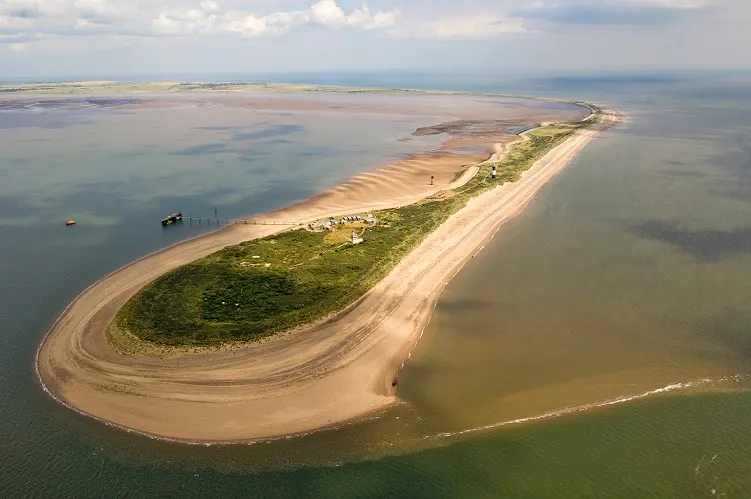 Aerial view of a curved sandy spit extending into shallow coastal waters with vegetation and small buildings along its length.
