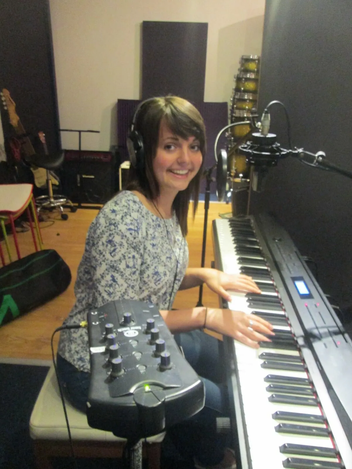 Young woman wearing headphones playing keyboard in a recording studio, smiling at the camera.