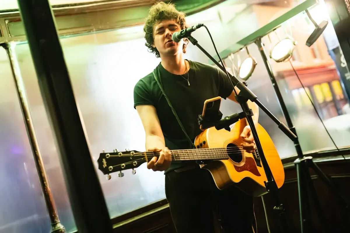 Young musician performing with acoustic guitar at a microphone in an indoor venue.