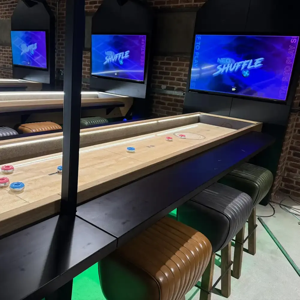 Shuffleboard tables with coloured pucks in an indoor gaming venue with screens displaying game graphics and leather seating.