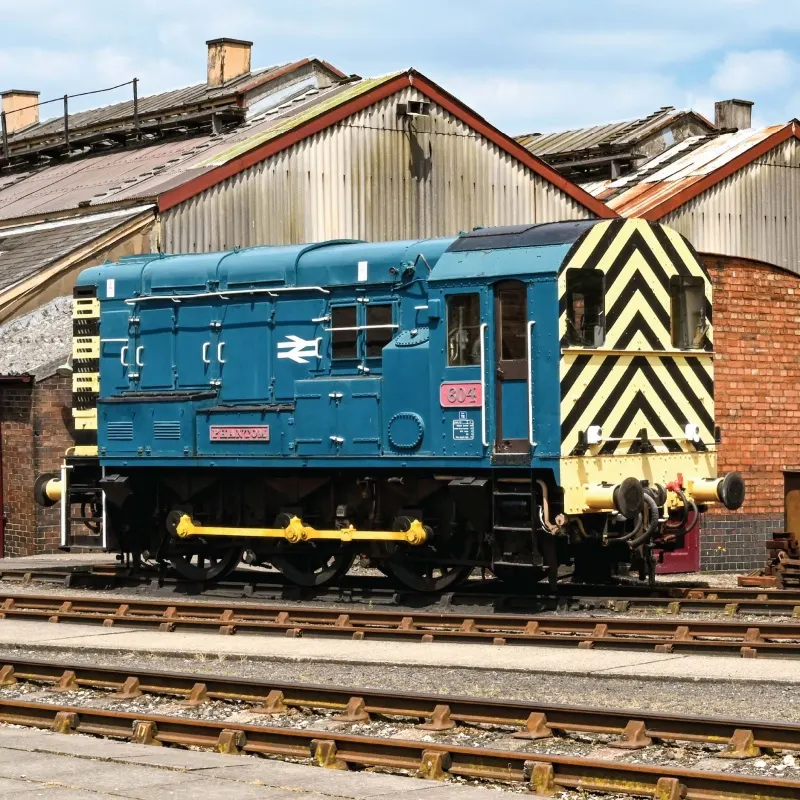 Blue diesel locomotive with yellow and black chevron marking on shed track at heritage railway depot.