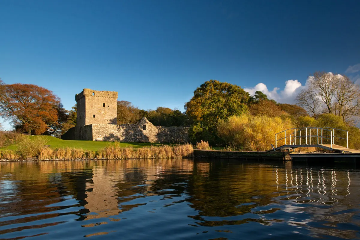 Stone castle tower beside a loch with autumn trees, footbridge and reedy shoreline under blue sky.