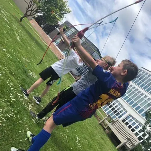 Two young people flying a kite together in a grassy field beside a brick building.