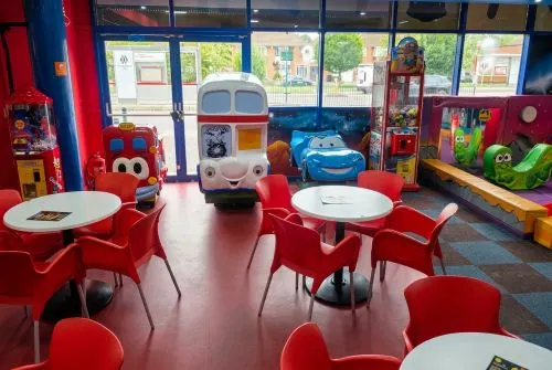 Colourful indoor play area with red tables and chairs, toy vehicles, and arcade games visible under large windows.