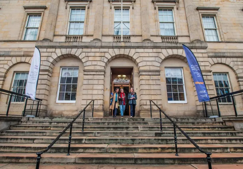 People standing at the entrance of a grand historic stone building with arched windows and central doorway.