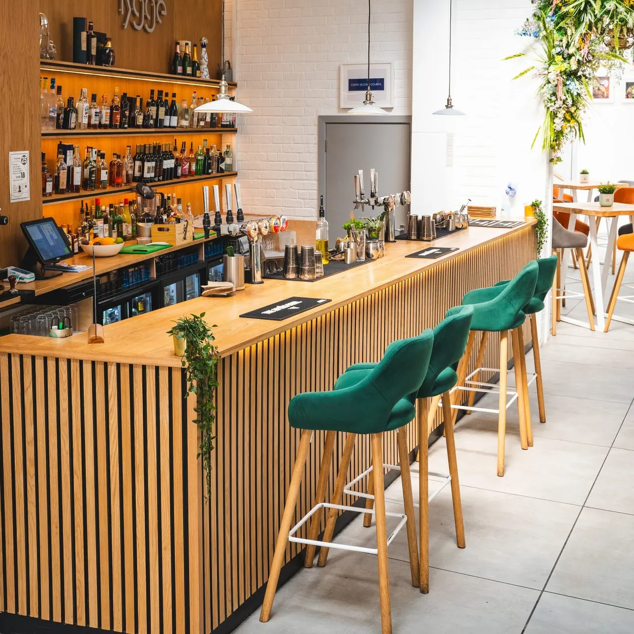 Modern bar interior with wooden counter, green bar stools, illuminated shelving displaying spirits, and plants.