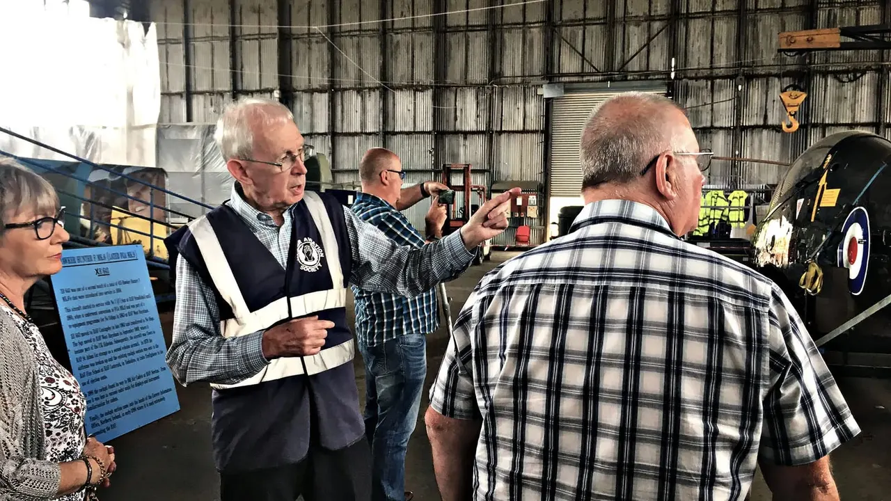 A tour guide in a high-visibility vest gestures whilst explaining vintage aircraft to a group of visitors inside a large hangar.