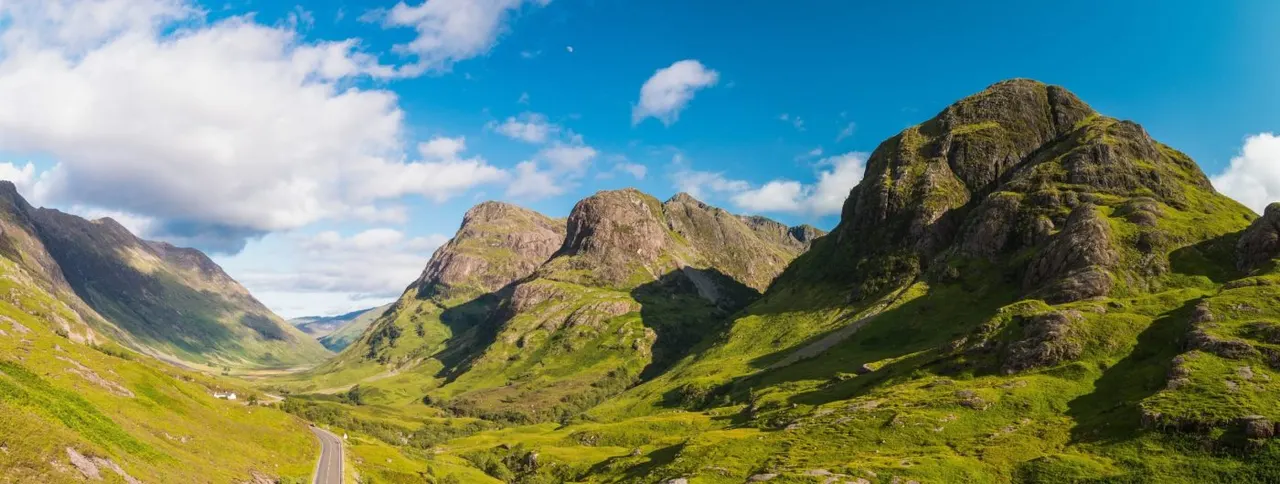 Highland valley landscape with rolling green hills, distinctive mountain peaks, winding road and scattered buildings under blue sky.
