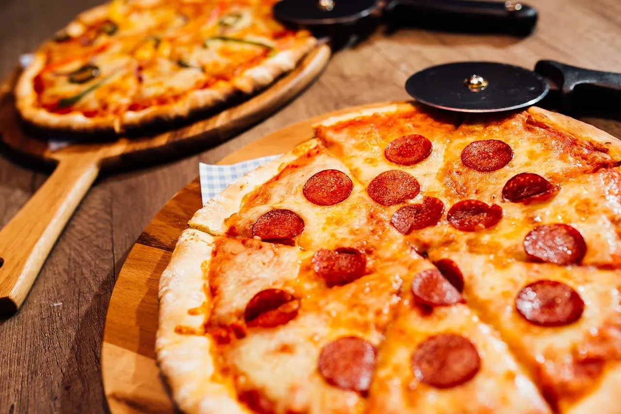 Two pepperoni pizzas on wooden boards with pizza cutters on a wooden table.