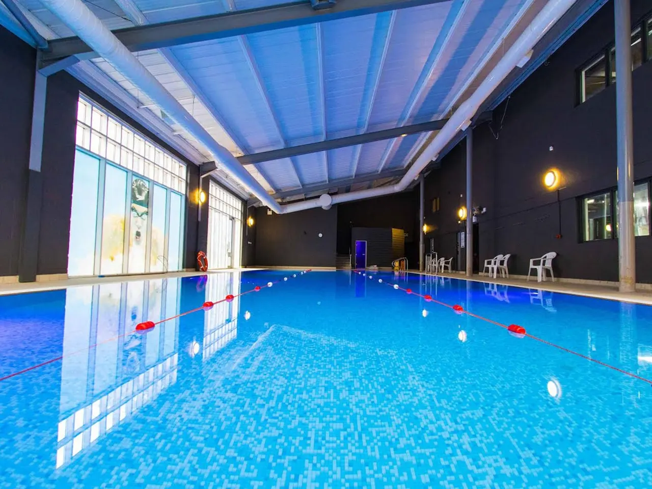Indoor swimming pool with lane dividers, lit by blue ceiling panels and wall lights, with white chairs and dark walls.
