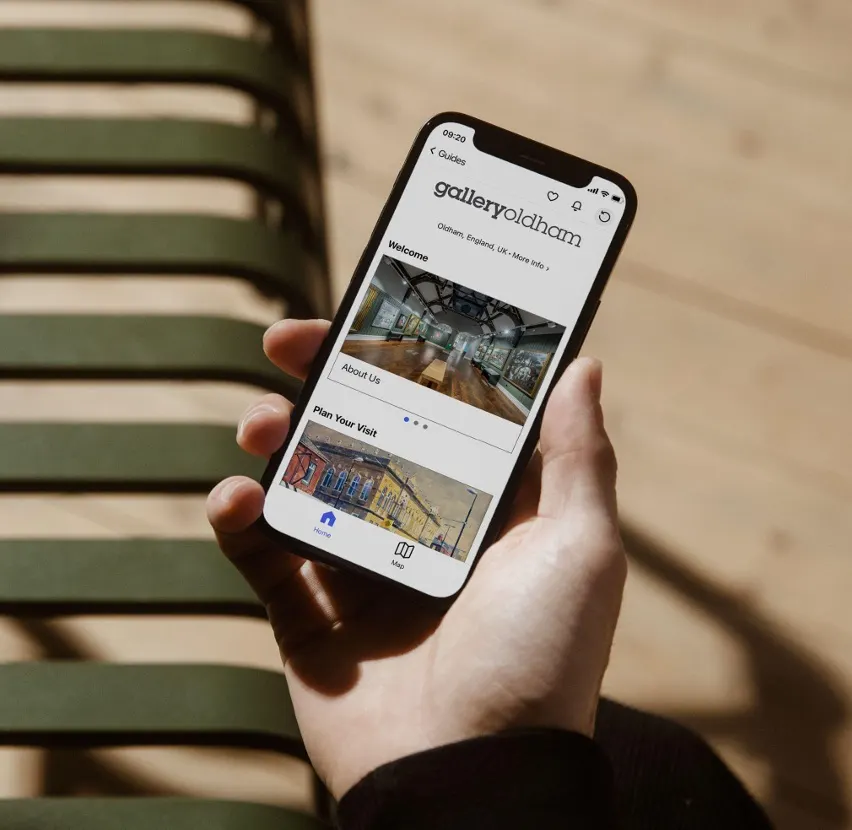 Person holding smartphone displaying Gallery Oldham website, sat on wooden bench outdoors.