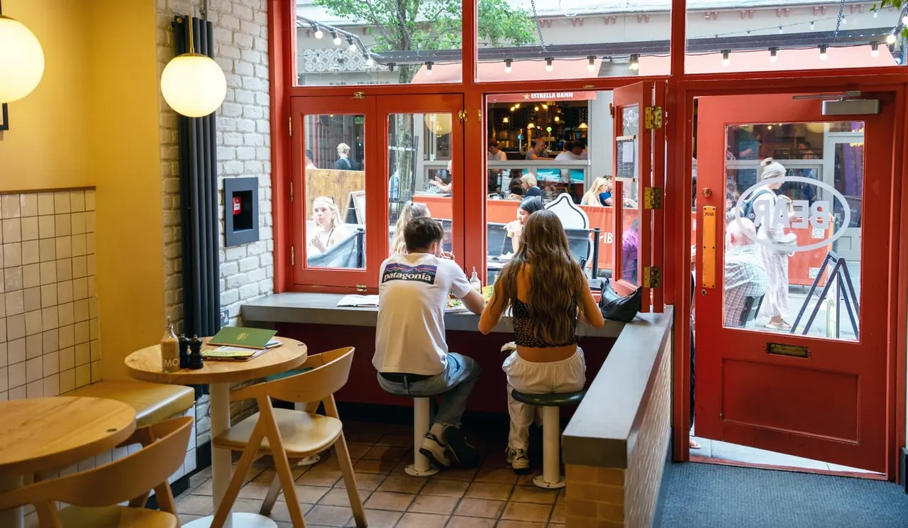 Two children sit at a window counter inside a colourful café, looking out at the street and other customers outside.