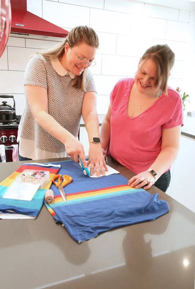 Two women decorating a blue fabric with rainbow stripes and craft materials on a kitchen counter.
