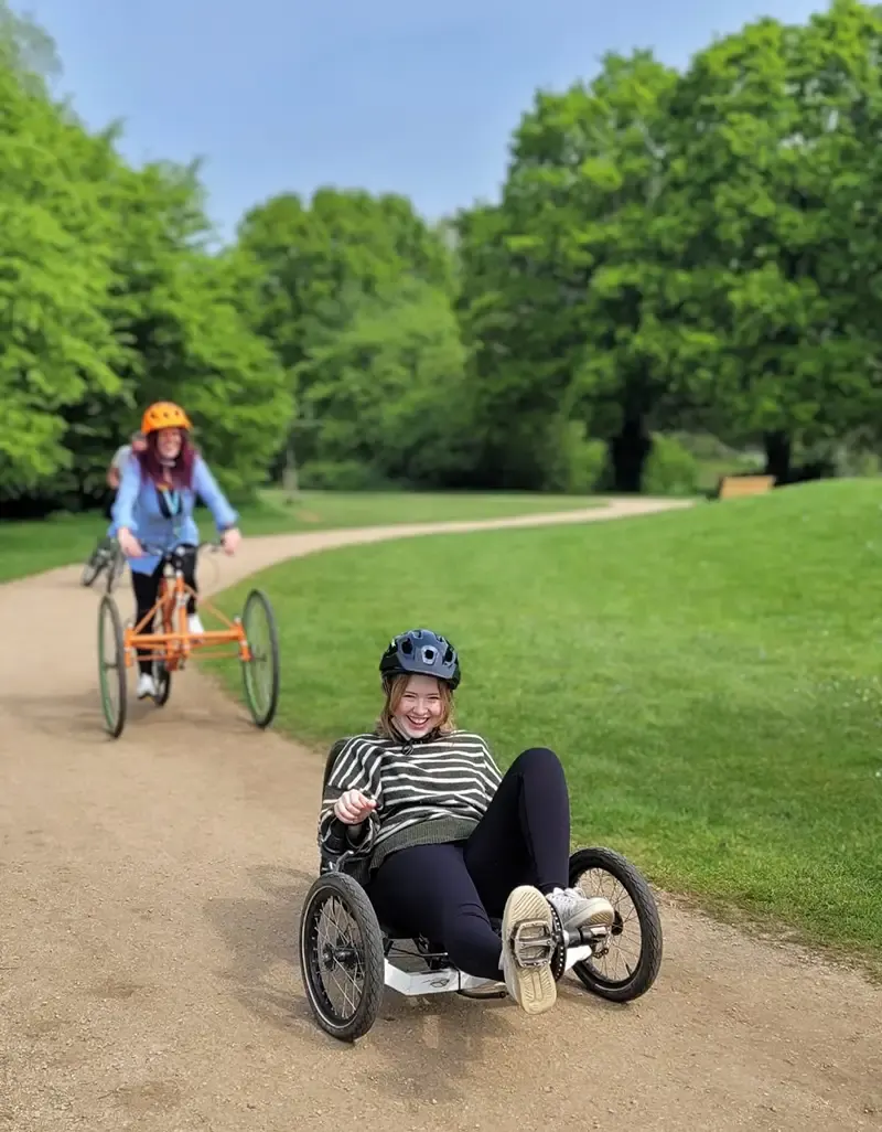 Two visitors riding recumbent tricycles on a park path bordered by green grass and trees on a sunny day.