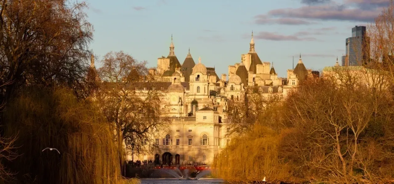 Ornate cream-coloured building with multiple golden domes viewed through bare winter trees lining a park pathway.