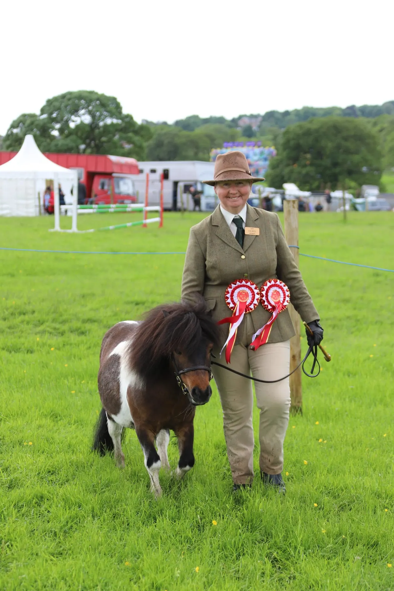 Woman in formal riding attire leading a small brown and white pony across a grass field at a country show.