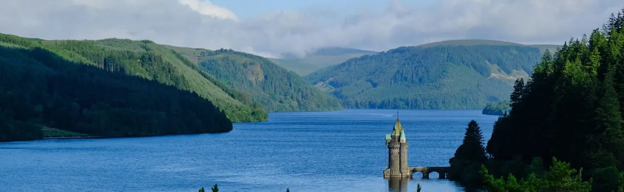A stone tower stands in a deep blue lake surrounded by forested hills under partly cloudy sky.