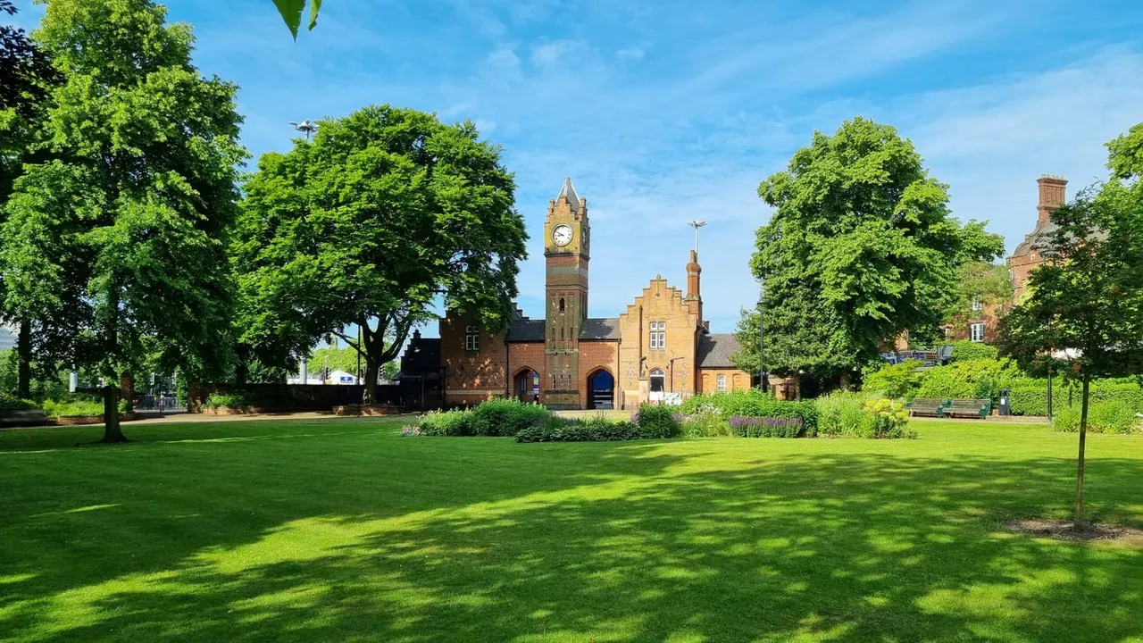 A sunlit park with expansive green lawns, mature trees, and a Victorian brick building with a clock tower in the background.