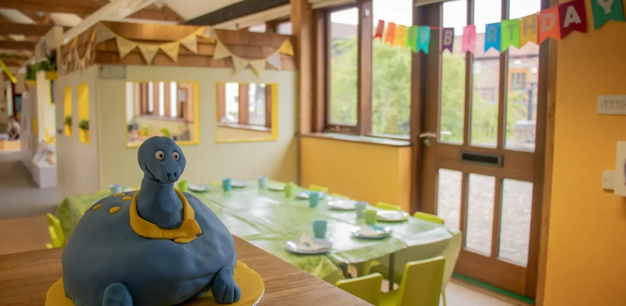 A blue toy character sits on a table in a bright children's party room with a set dining table, colourful bunting and large windows.