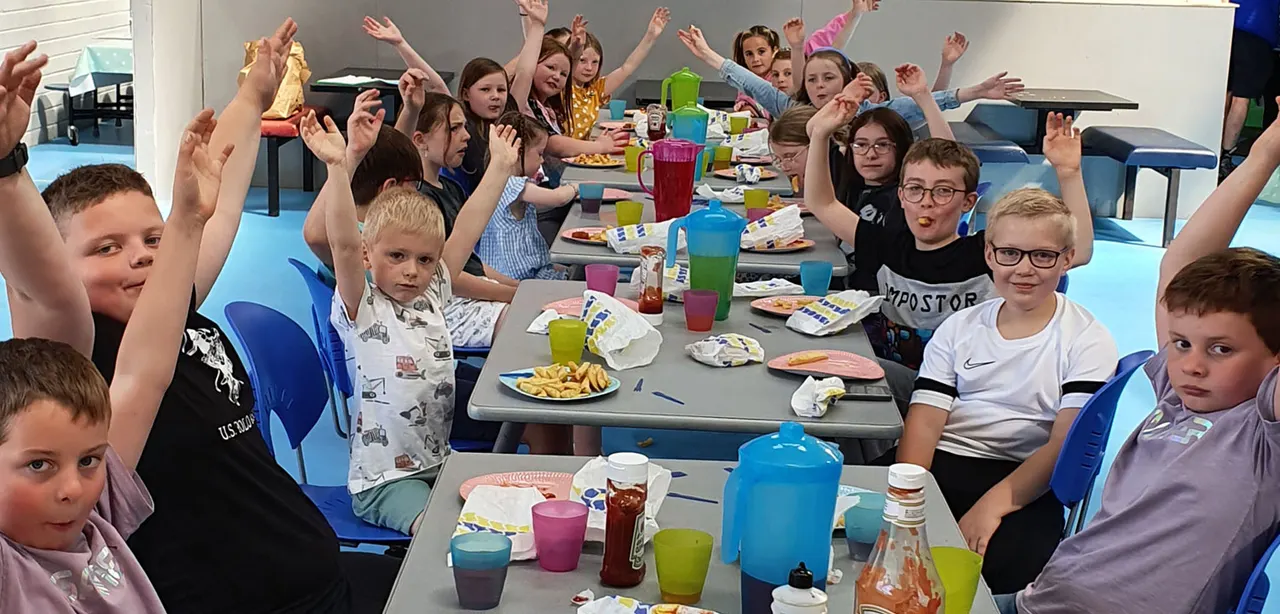 Group of children seated at long tables with food and drinks, many with hands raised in celebration.