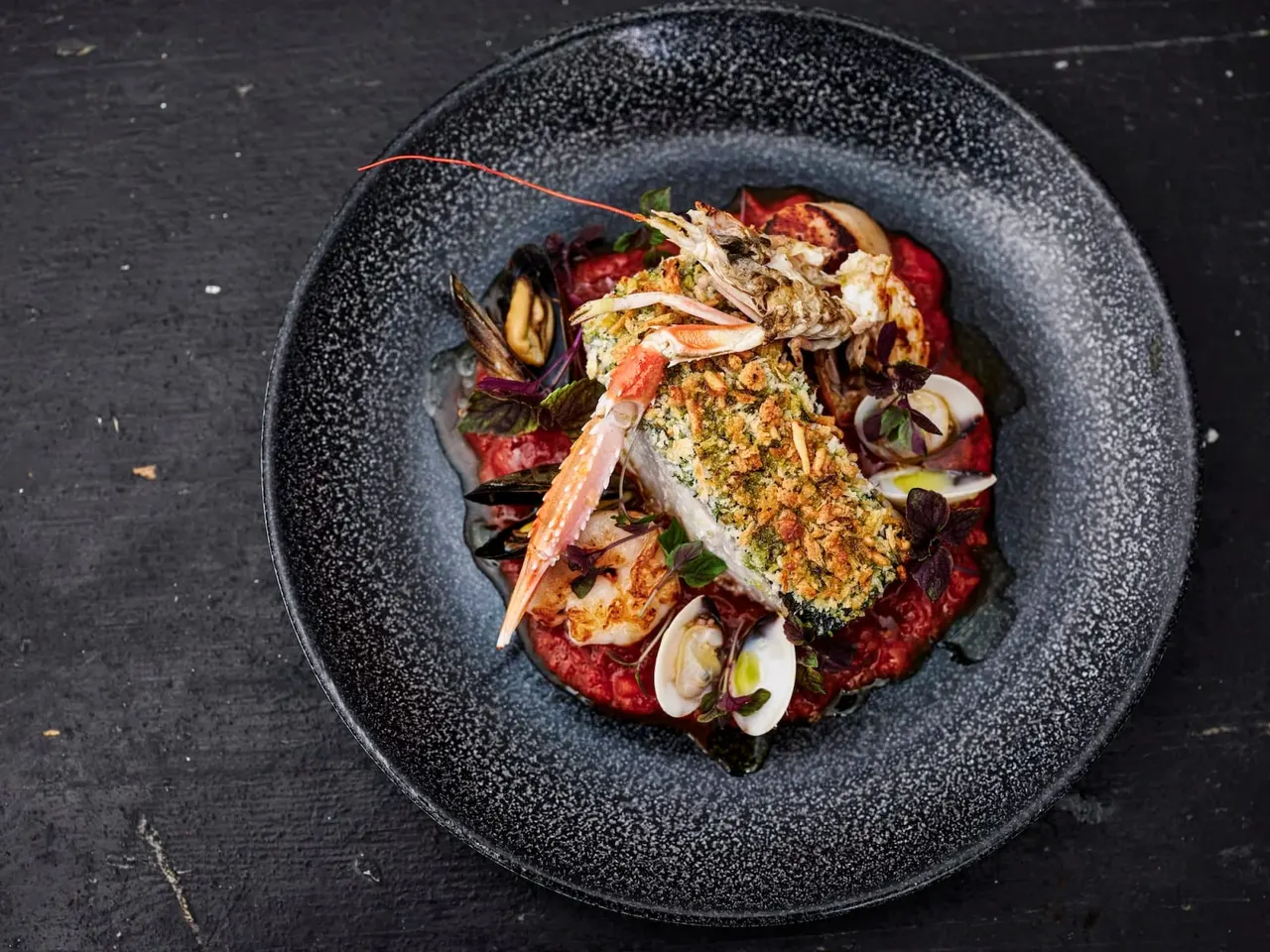 Plated seafood dish with crab, prawns, clams and herb crust on red sauce in dark bowl.