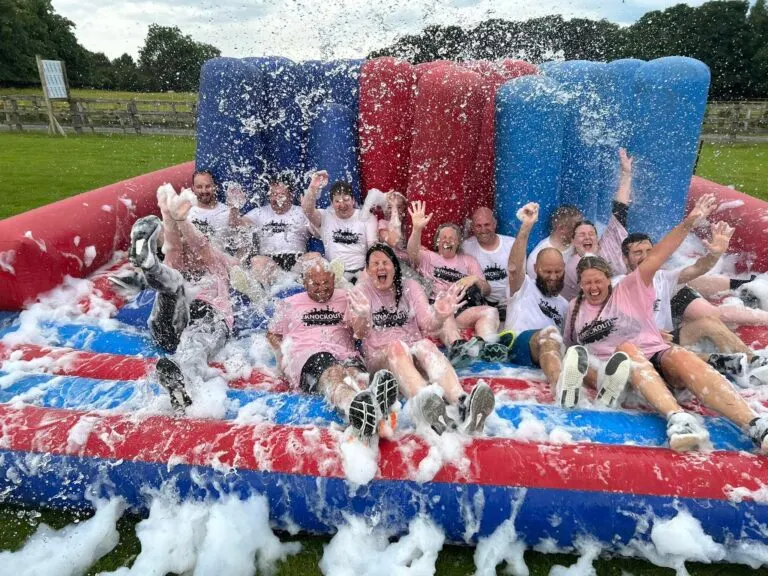 Group of people enjoying a foam and soap slide activity on a red, white and blue inflatable structure in a grassy field.