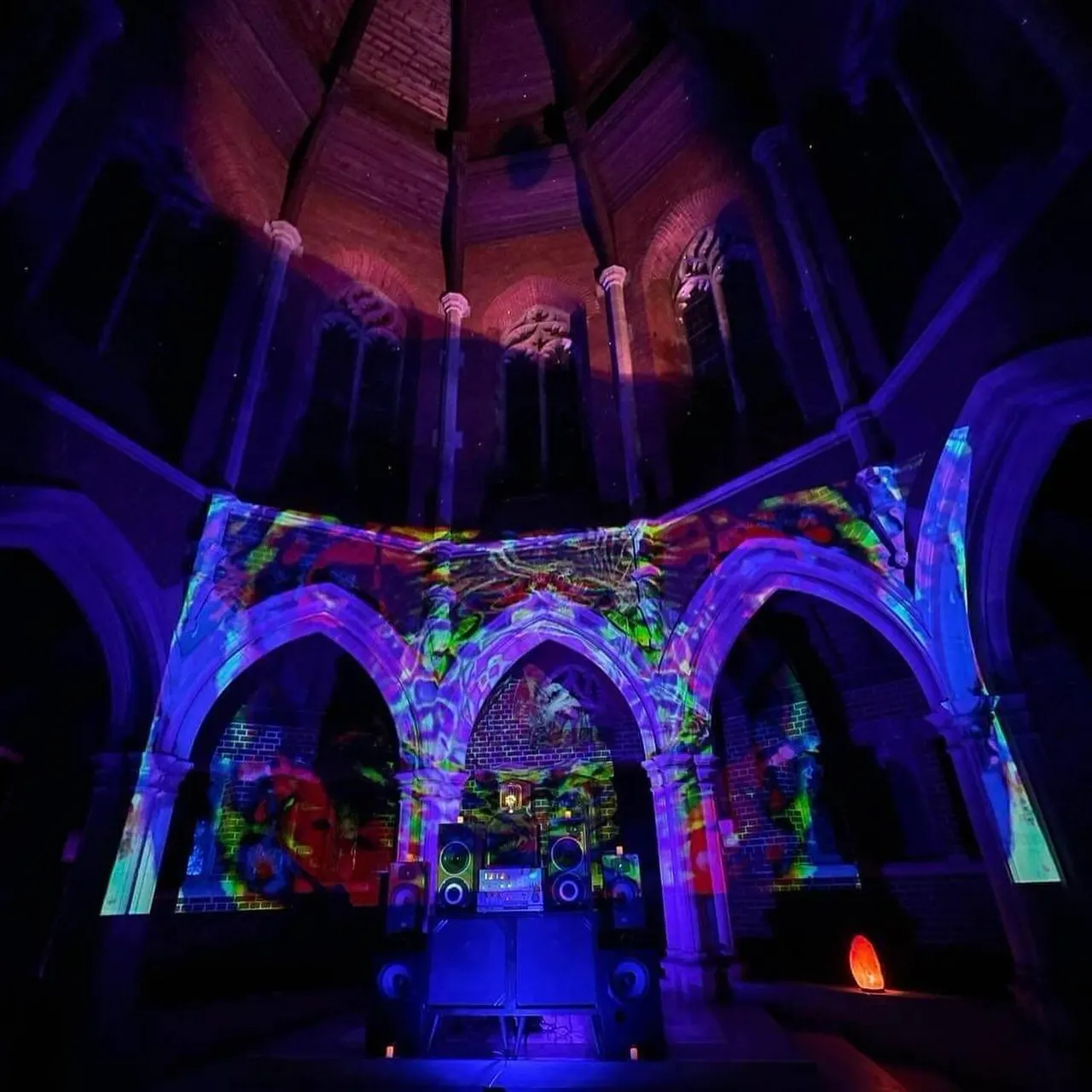Historic stone church interior with Gothic arches illuminated in vibrant purple, blue and multicoloured lighting, featuring a DJ booth and sound system.