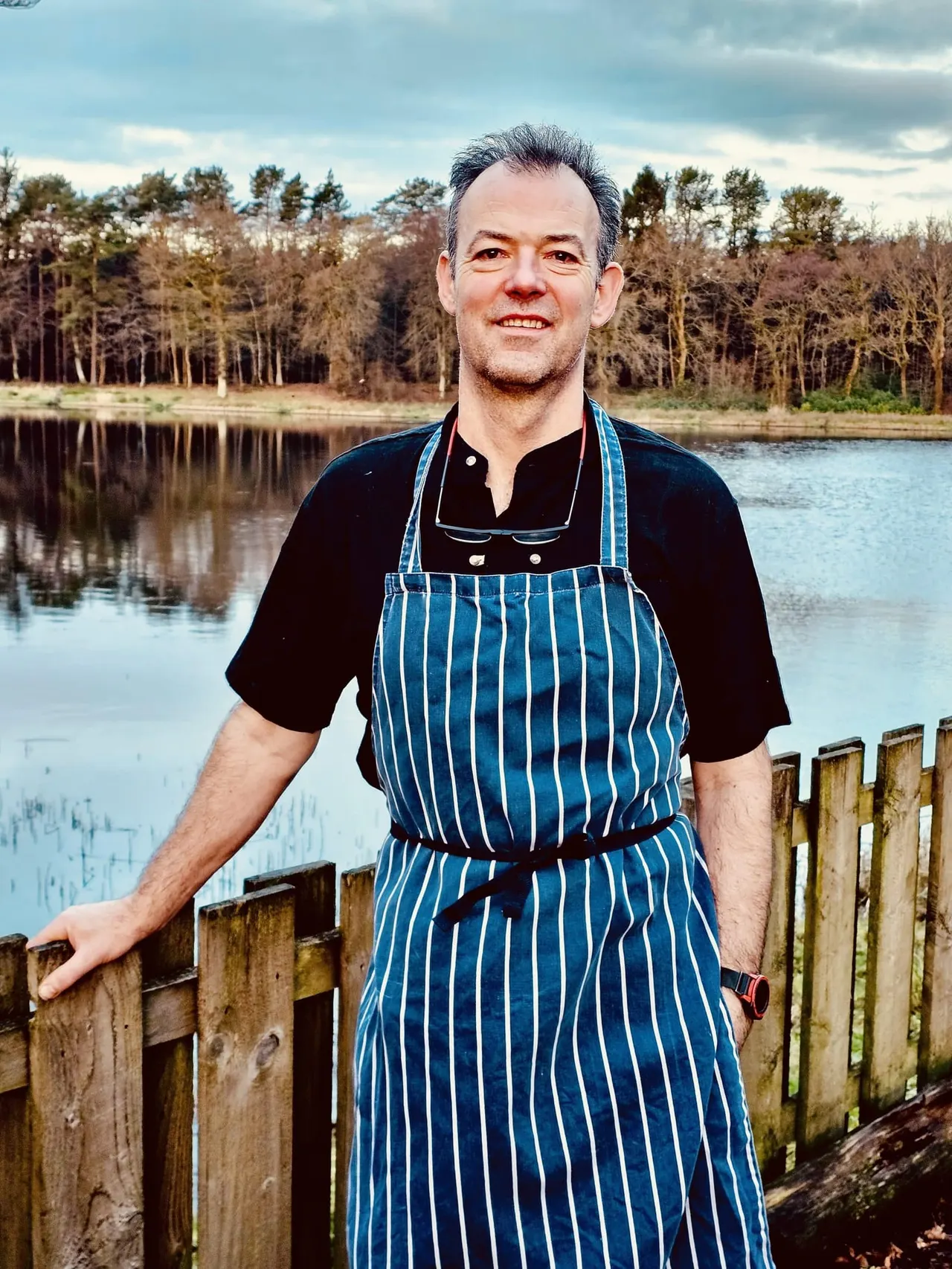 Man in blue striped apron standing by a wooden fence beside a still loch with bare trees beyond.
