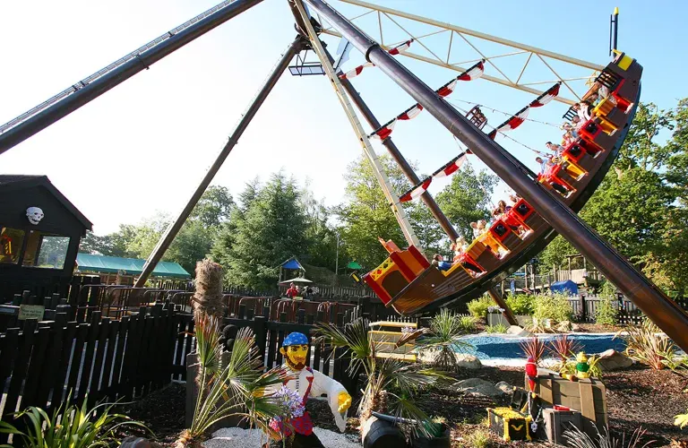 A red and yellow roller coaster ride tilted at a steep angle against a blue sky, with tropical plants and wooden structures below.