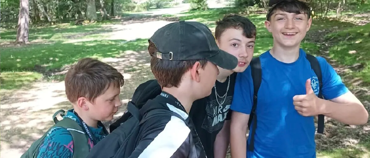 Four boys smiling at the camera on a woodland path during a daytime outdoor activity.