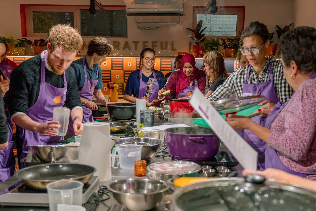 Group of people wearing purple aprons cooking together in a bright kitchen classroom with pots, pans and ingredients on the counter.