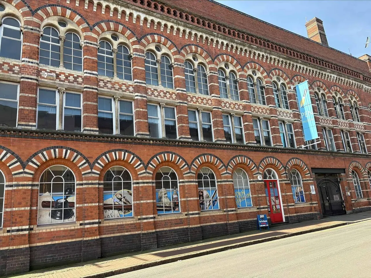 Victorian red-brick building with decorative arched windows and a blue banner, housing the Pen Museum.