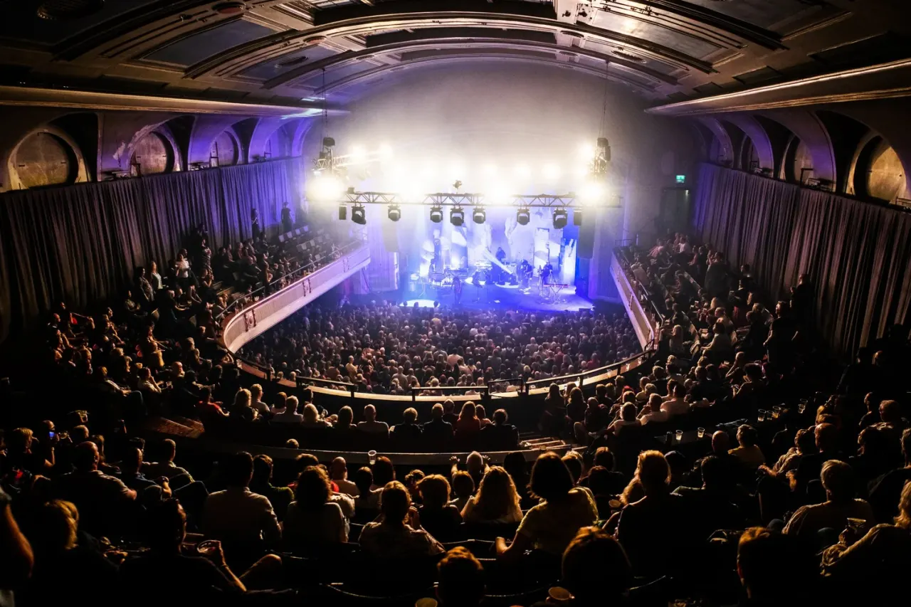 A packed concert hall with a large crowd watching a live band perform on stage under bright lights.