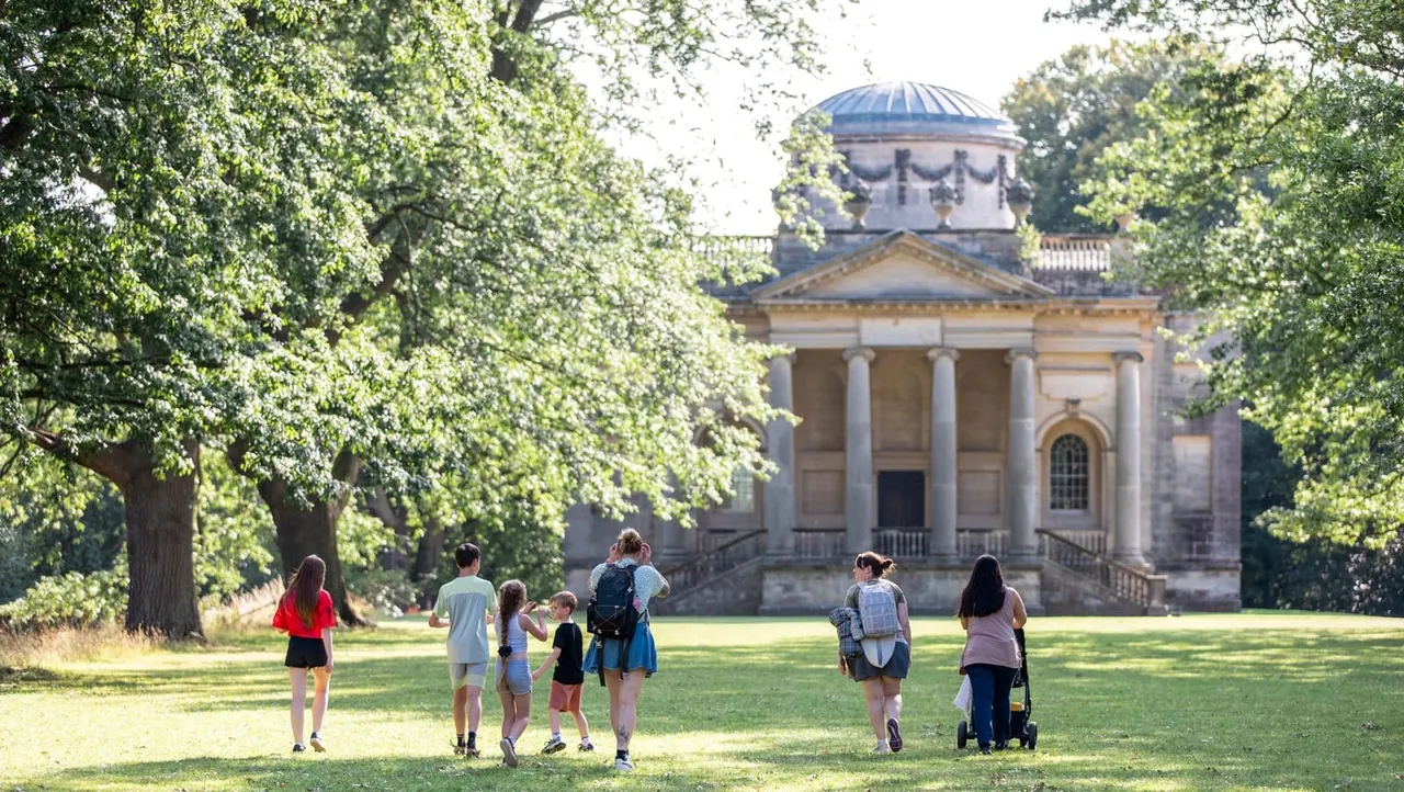Children and adults enjoying a grassy parkland with mature trees, a neoclassical chapel with blue dome visible in the background.