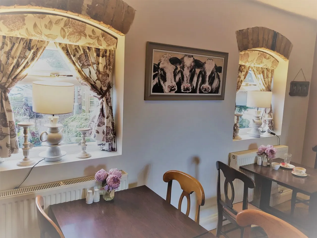 Interior of a farmhouse dining room with framed cow artwork on wall, patterned curtains, wooden tables and chairs, fresh flowers, and table lamps.