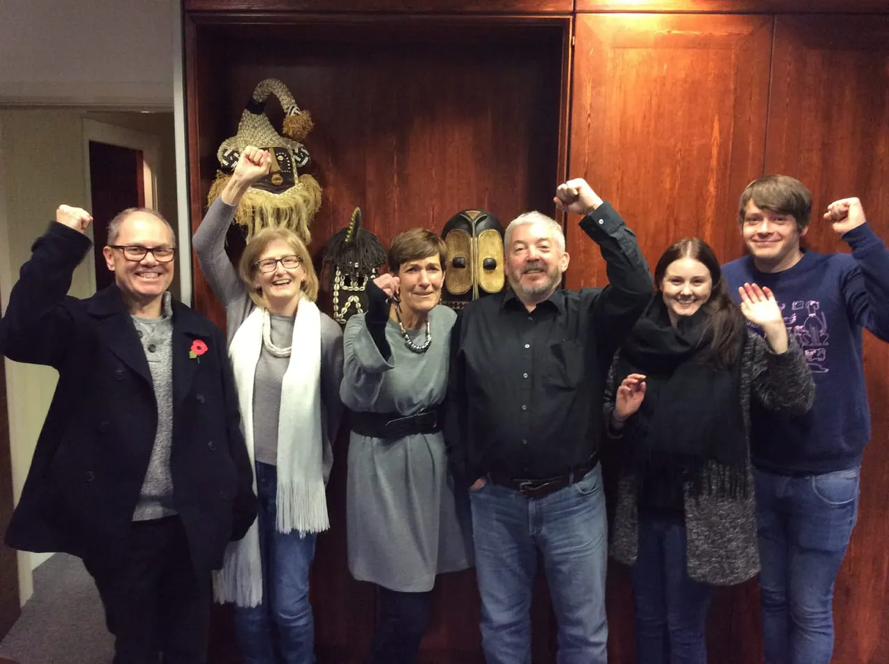 Group of six adults posing cheerfully in front of wooden wall display with decorative masks and wizard figurine.