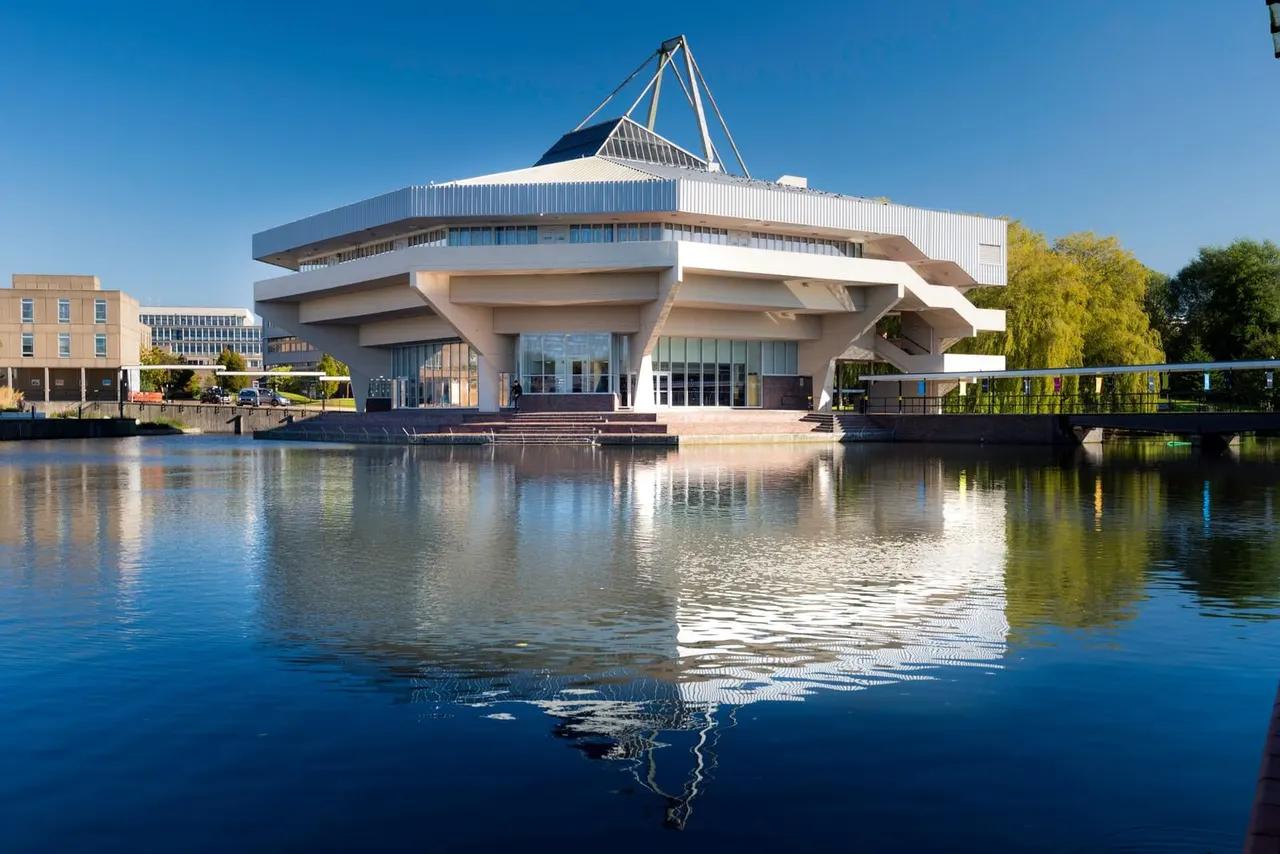 Modern waterfront building with distinctive white curved architecture and sail-like roof structure, reflected in calm water on a clear day.
