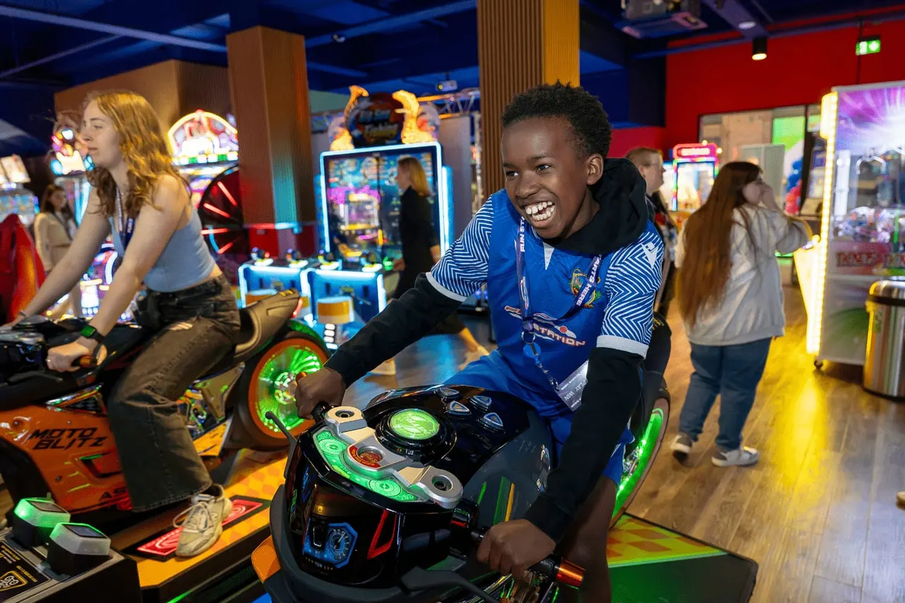 Young person playing on a racing arcade game in a brightly lit amusement arcade with other visitors and gaming machines in the background.