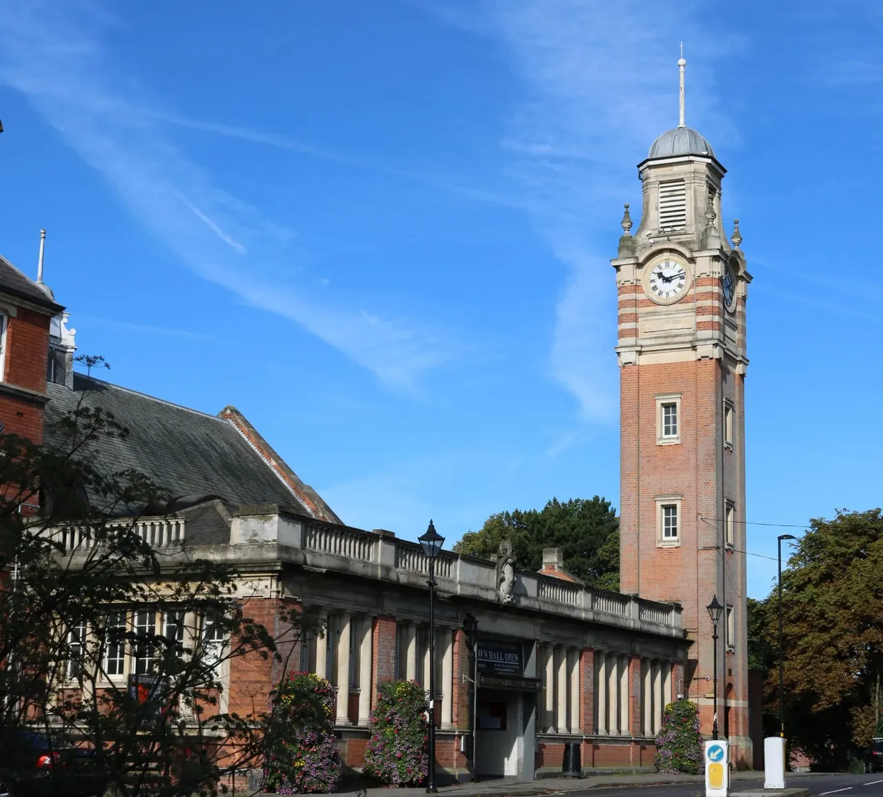 A red brick Victorian town hall with a prominent clock tower against a clear blue sky, surrounded by trees and flowering planters.