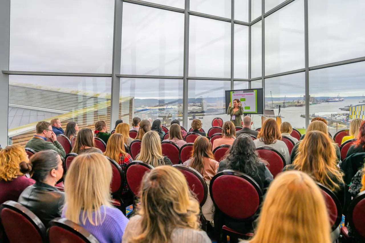 Audience seated in red chairs watching a presentation in a high-rise venue with expansive views of a coastal city, harbour and mountains.