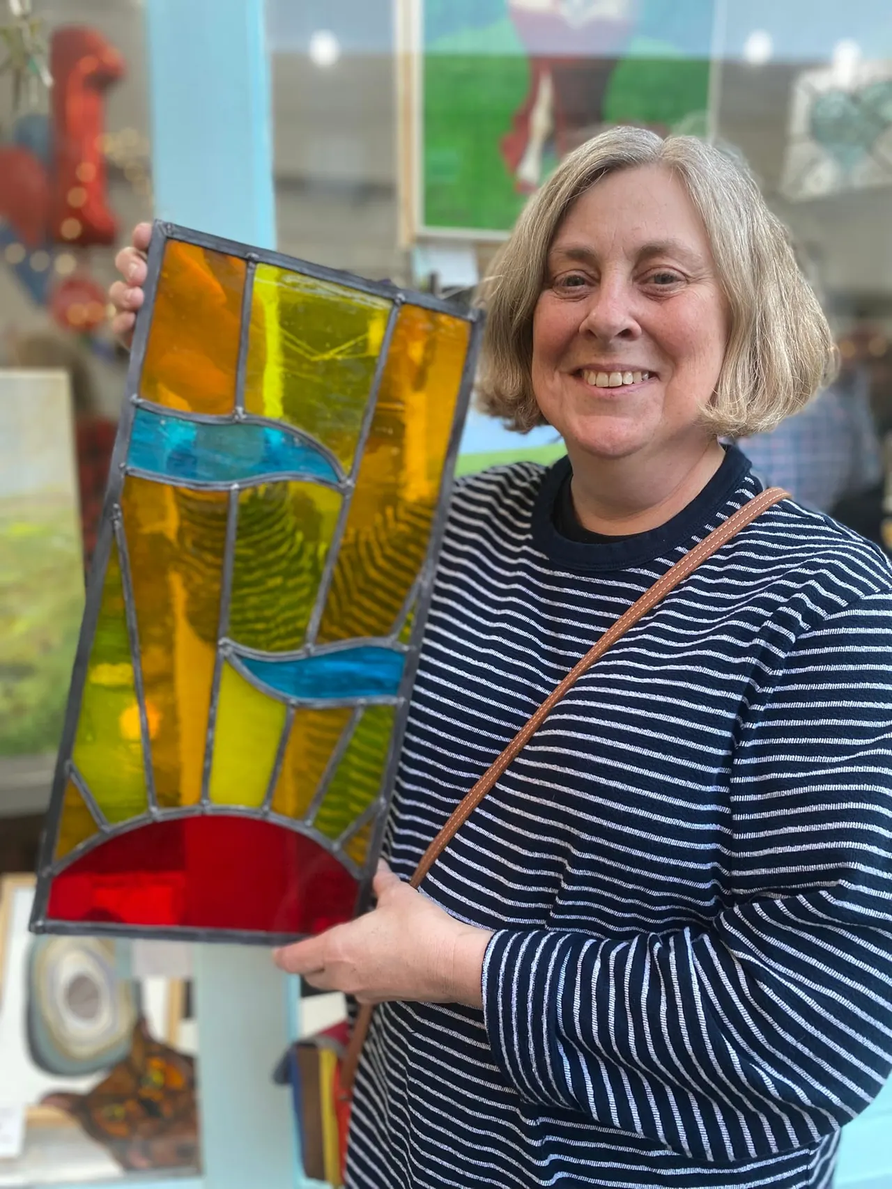 Woman holding a colourful stained glass panel inside an art gallery or studio space.