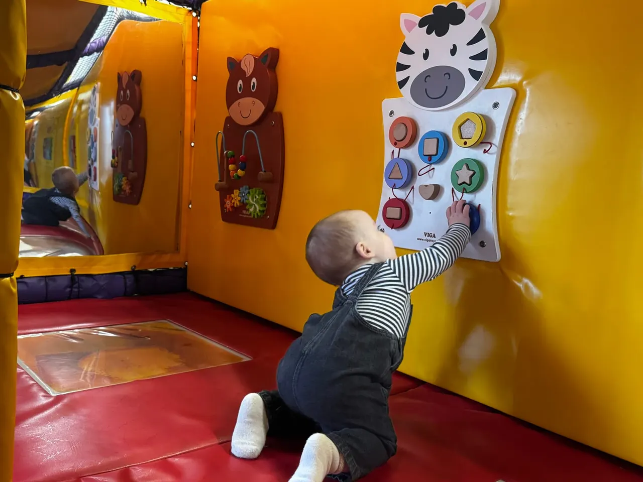 A toddler reaches up to press buttons on an interactive wall panel featuring a zebra design inside a soft play area with colourful padded walls.
