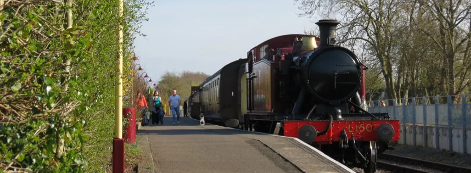 A heritage steam locomotive with red and black livery stands at a railway platform with passengers boarding nearby.